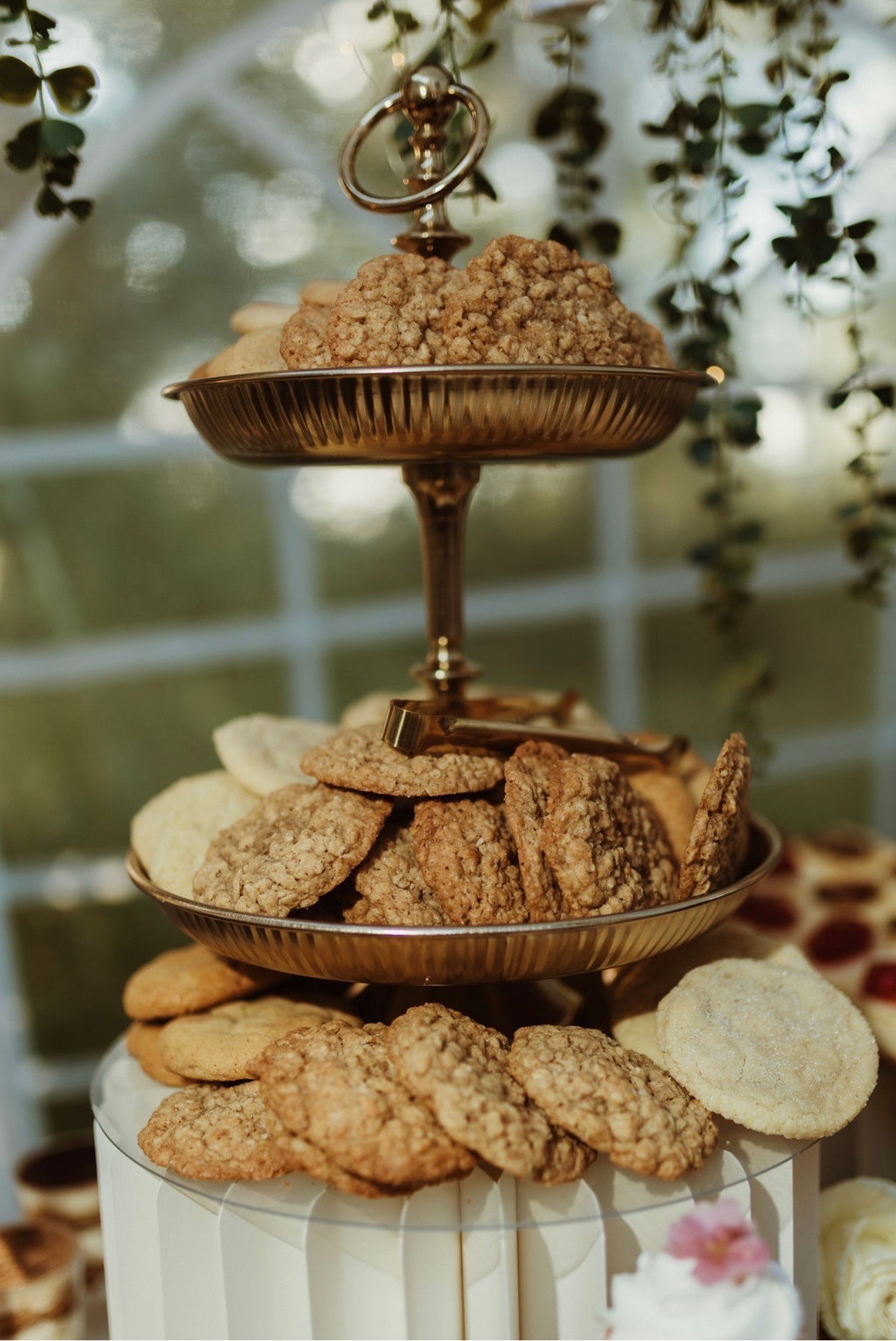 A gold two-tier stand piled with oatmeal and sugar cookies, gold tongs resting on top