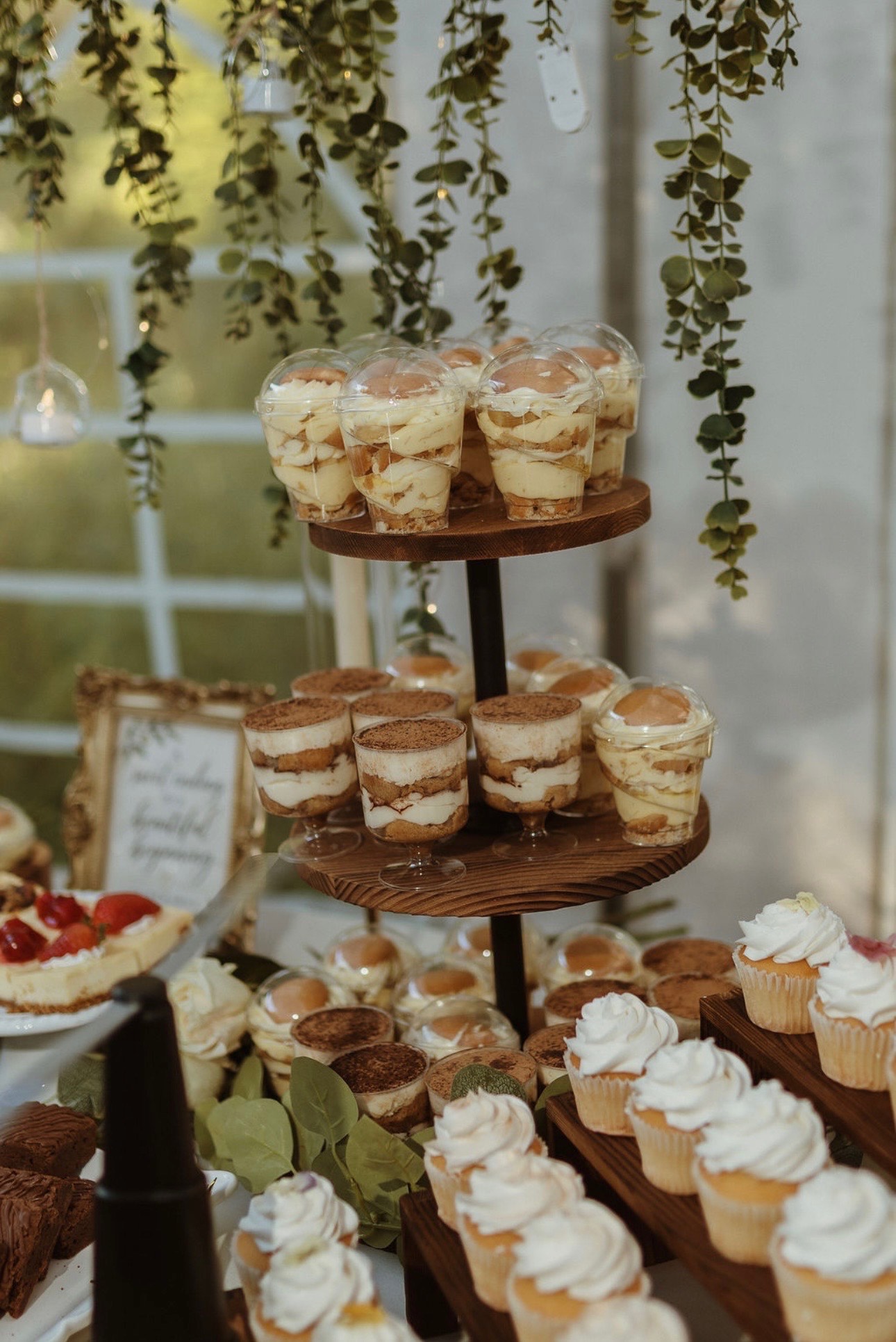 A tiered stand of banana pudding and tiramisu cups with frosted cupcakes beneath hanging eucalyptus